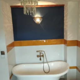 Biała wanna na złotych nóżkach w łazience z ciemnoniebieską ścianą, oświetlona ozdobnym żyrandolem i kinkietem. Żółte płytki stanowią akcent kolorystyczny.