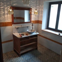 Oleh Tuz EUROBILD - Wykończona łazienka z drewnianą szafką pod umywalką, wzorzystą podłogą i oknem. Widoczne lampy wiszące i akcesoria. Całość w stylu retro.