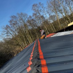 Oleh Tuz EUROBILD - Pracownik montuje membranę dachową na szarym dachu z blachy na tle drzew i błękitnego nieba. Widok z góry, skupienie na detalu łączenia membrany.