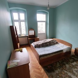 Oleh Tuz EUROBILD - Pokój z łóżkiem, komodą i toaletką w odcieniach błękitu i brązu. Parkiet na podłodze, okna z białymi ramami. W tle drzwi balkonowe i wisząca żarówka.