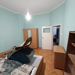 Oleh Tuz EUROBILD - Pokój przed remontem: parkiet, błękitne ściany, drewniane meble, łóżko z narzutą i lampa. Widok na otwarte drzwi do kolejnego pomieszczenia.