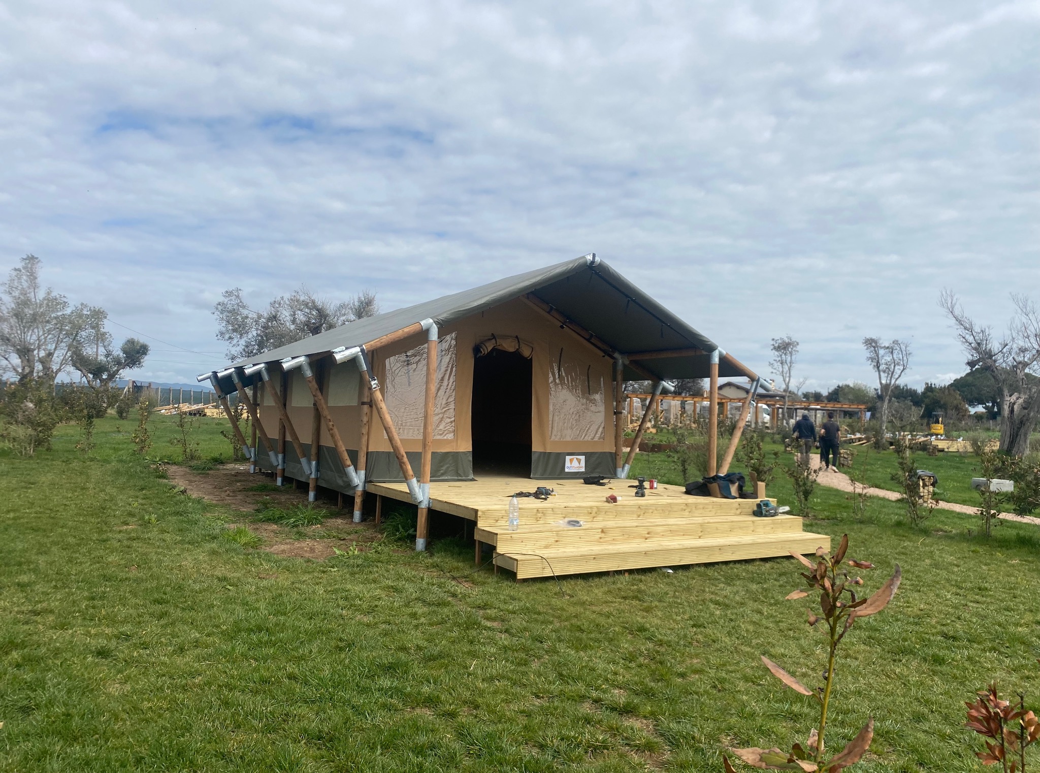 Wzniesiona konstrukcja namiotowa z drewnianym podestem na zielonej trawie. Widoczne narzędzia i elementy montażowe, sugerujące etap budowy w otoczeniu drzew i błękitnego nieba.