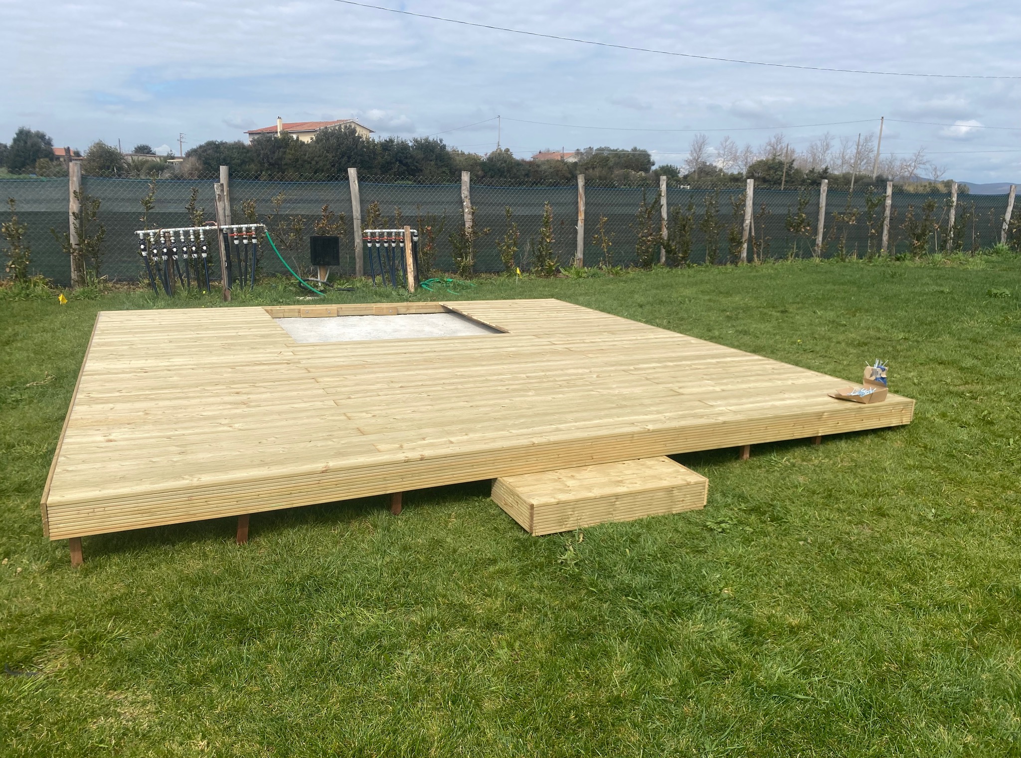 Nowy taras z jasnego drewna na trawie, z wycięciem na basen. W tle siatka ogrodzeniowa i zieleń. Widoczne narzędzia i akcesoria montażowe.