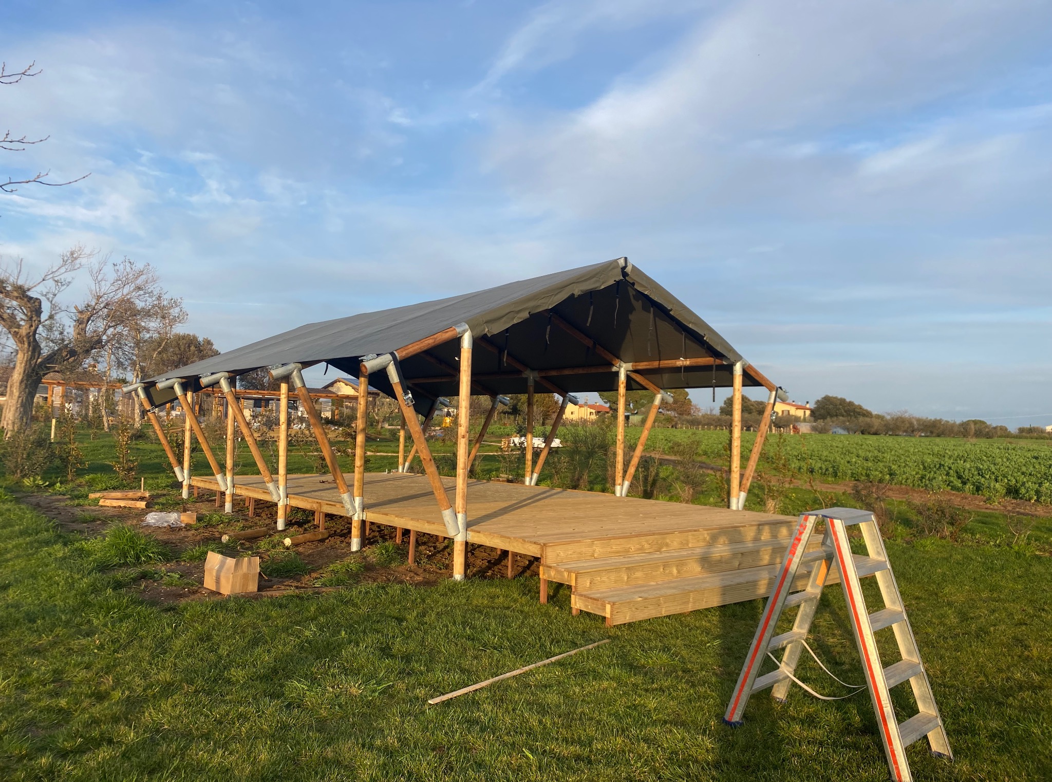 Nowoczesna altana z drewnianą podłogą i czarnym zadaszeniem w trakcie budowy. Na pierwszym planie drabina, w tle zielone pole i błękitne niebo.