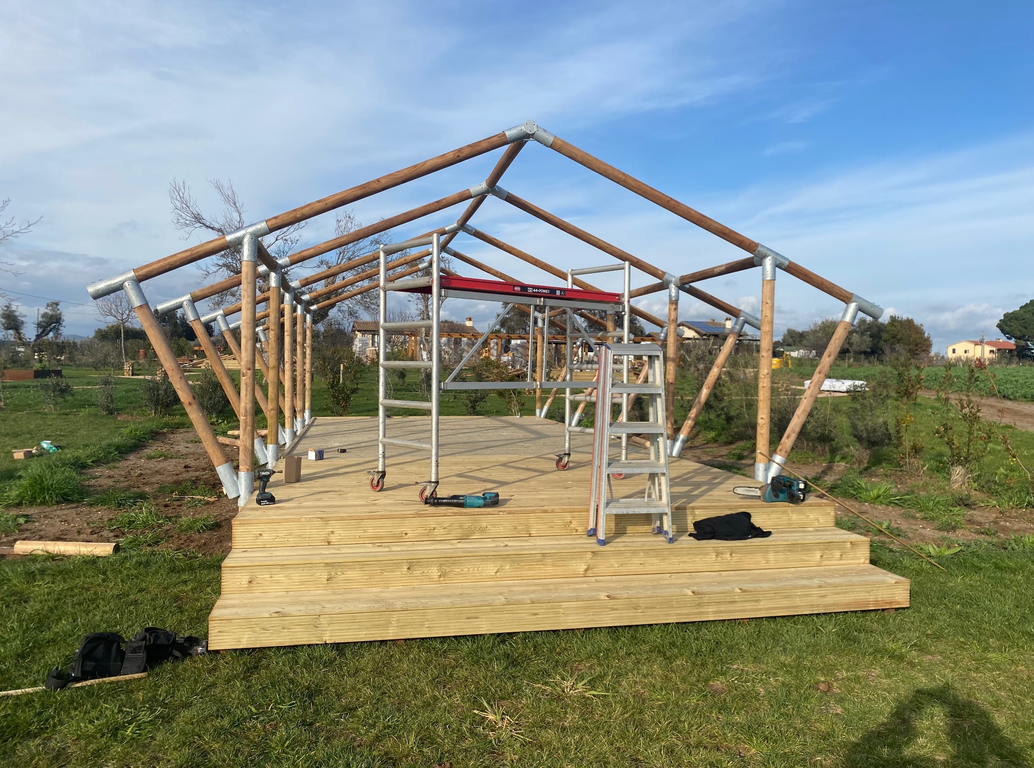 Konstrukcja altany w trakcie budowy: drewniany szkielet z metalowymi złączkami na drewnianym podeście. Widoczne narzędzia i rusztowanie.