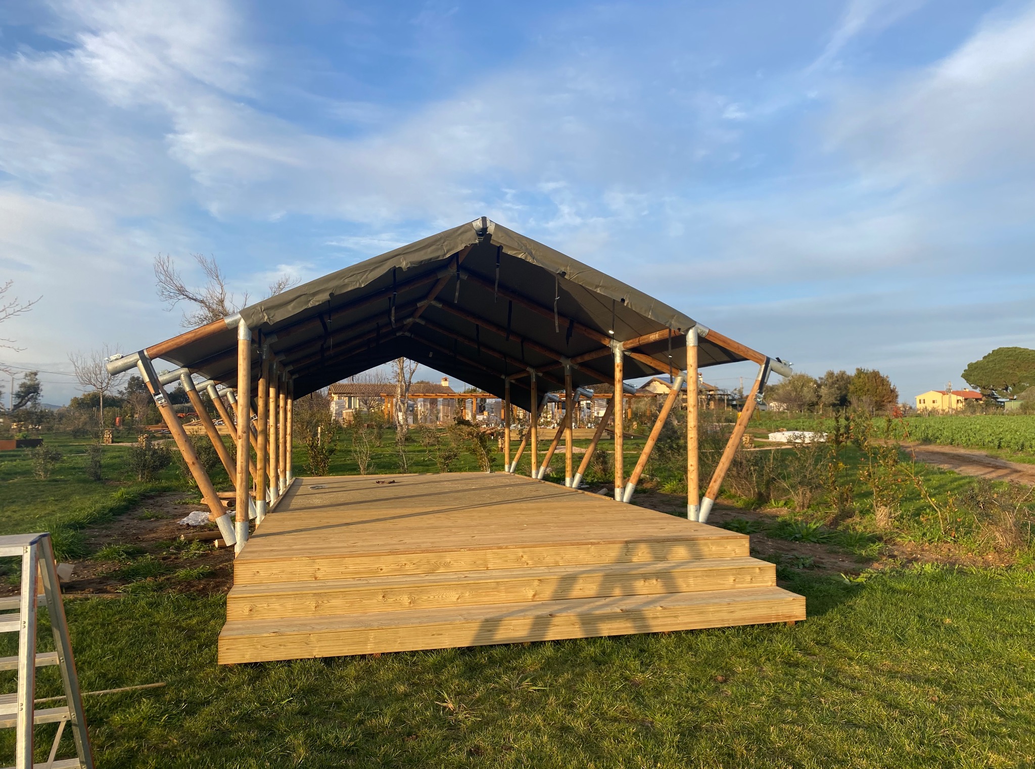 Drewniana konstrukcja z zadaszeniem na trawniku, widok z dołu. Schody prowadzą na podest. W tle domy i błękitne niebo. Częściowo widoczna drabina.