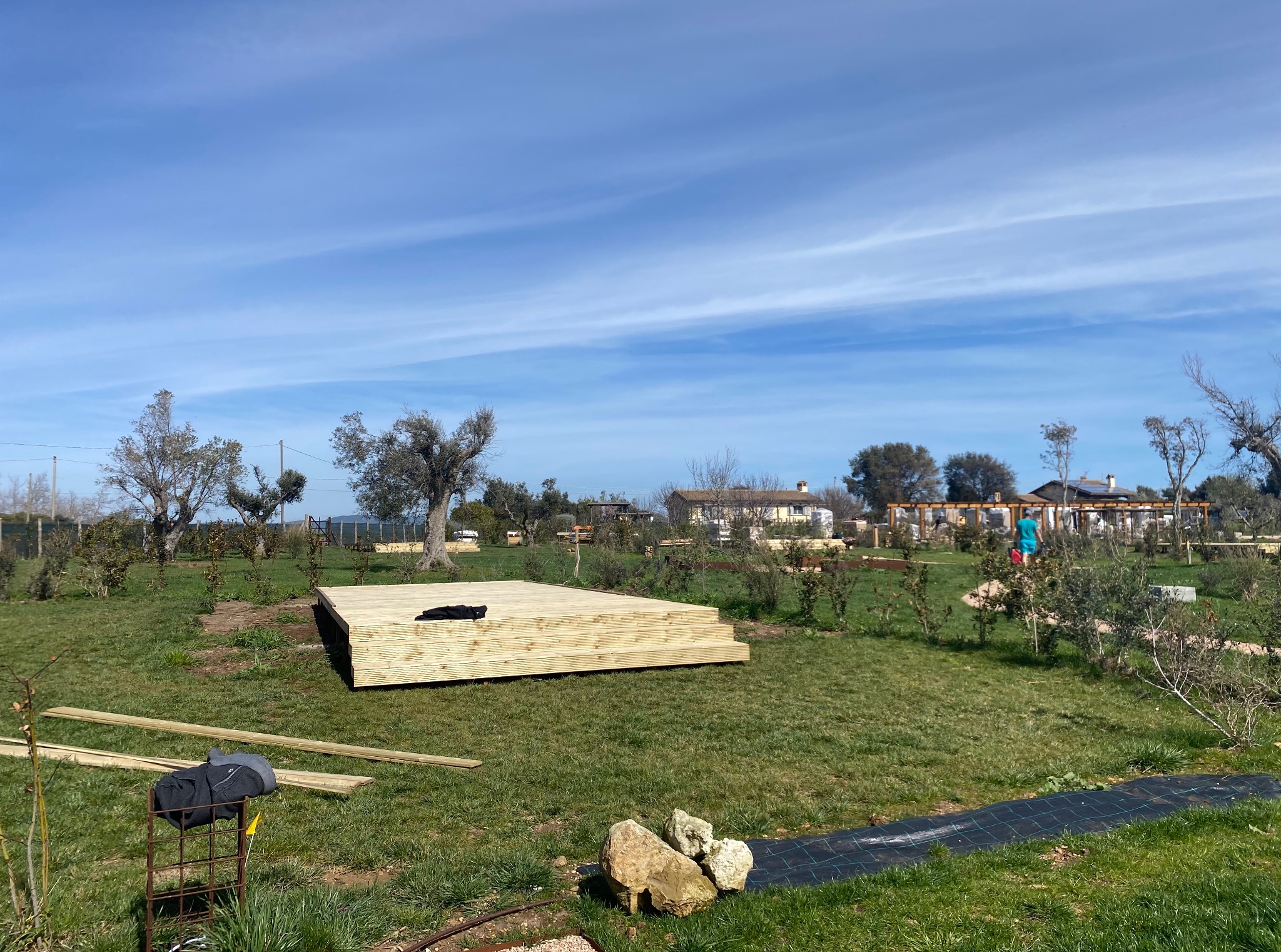 Drewniany podest tarasu w trakcie budowy na zielonej trawie, z elementami narzędzi i roślinności w tle, pod błękitnym niebem. Widoczne deski i elementy konstrukcyjne.