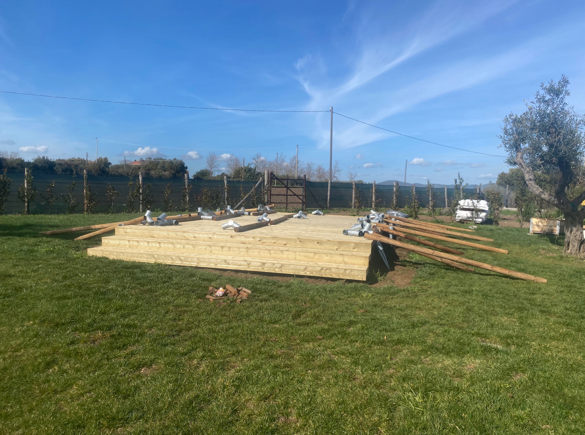 Drewniany taras w trakcie budowy z elementami konstrukcyjnymi i metalowymi łącznikami na trawniku, w tle ogrodzenie i drzewa pod błękitnym niebem.