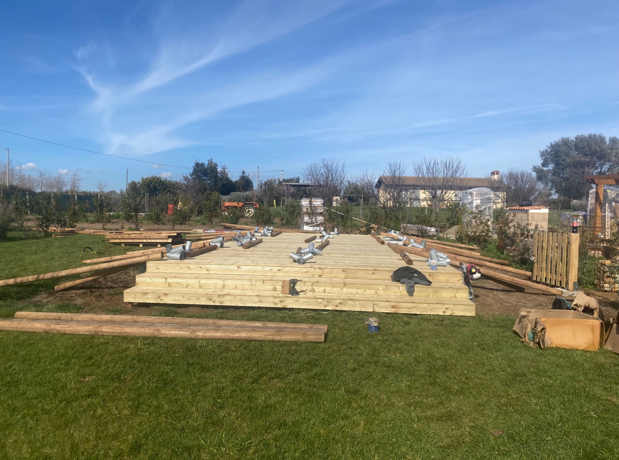 Budowa tarasu z drewna na trawie w słoneczny dzień. Widoczne deski, belki i elementy montażowe. Częściowo ukończona konstrukcja z widocznym tłem ogrodu.