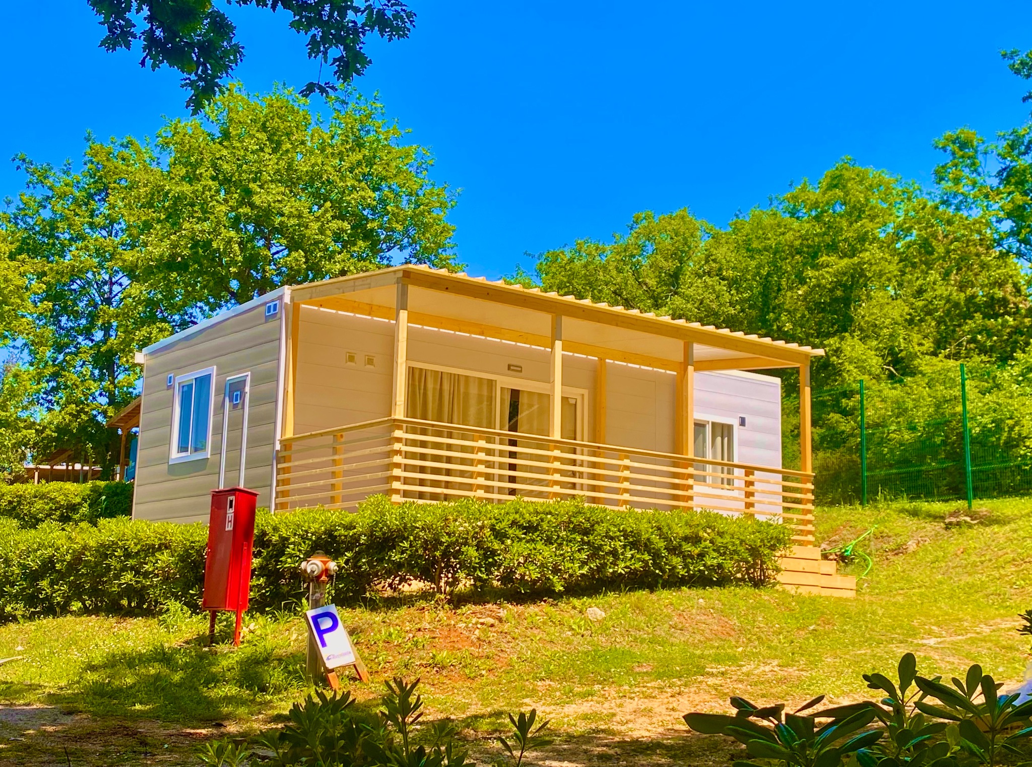 Nowoczesny, parterowy domek letniskowy z drewnianą konstrukcją zadaszenia, otoczony zielenią i błękitnym niebem. Widoczny hydrant i znak parkingu.