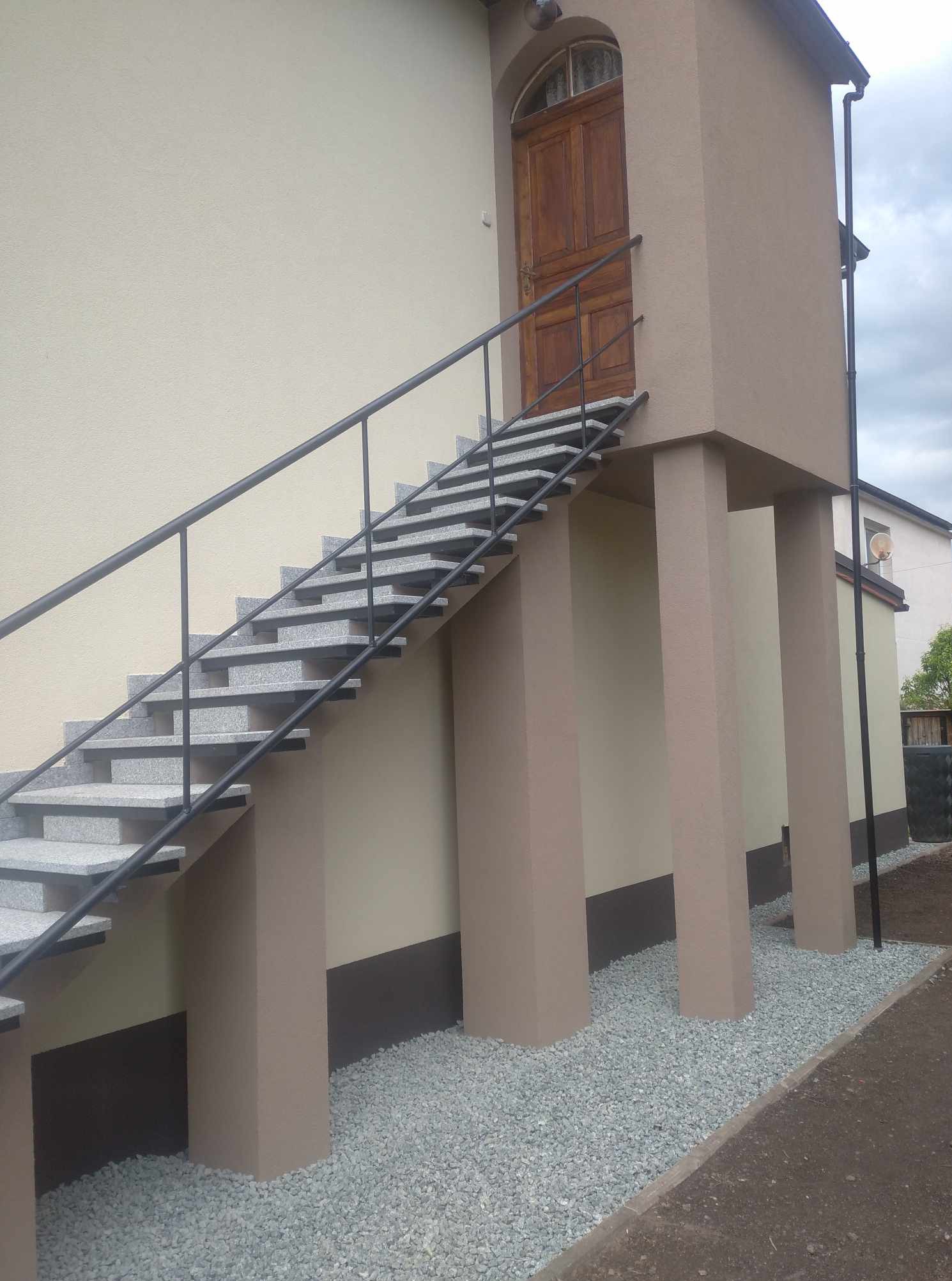 Zewnętrzne schody z szarego granitu z czarną balustradą, prowadzące do drewnianych drzwi na podwyższeniu, wsparte na beżowych kolumnach. Otoczenie wyłożone drobnym grysem.
