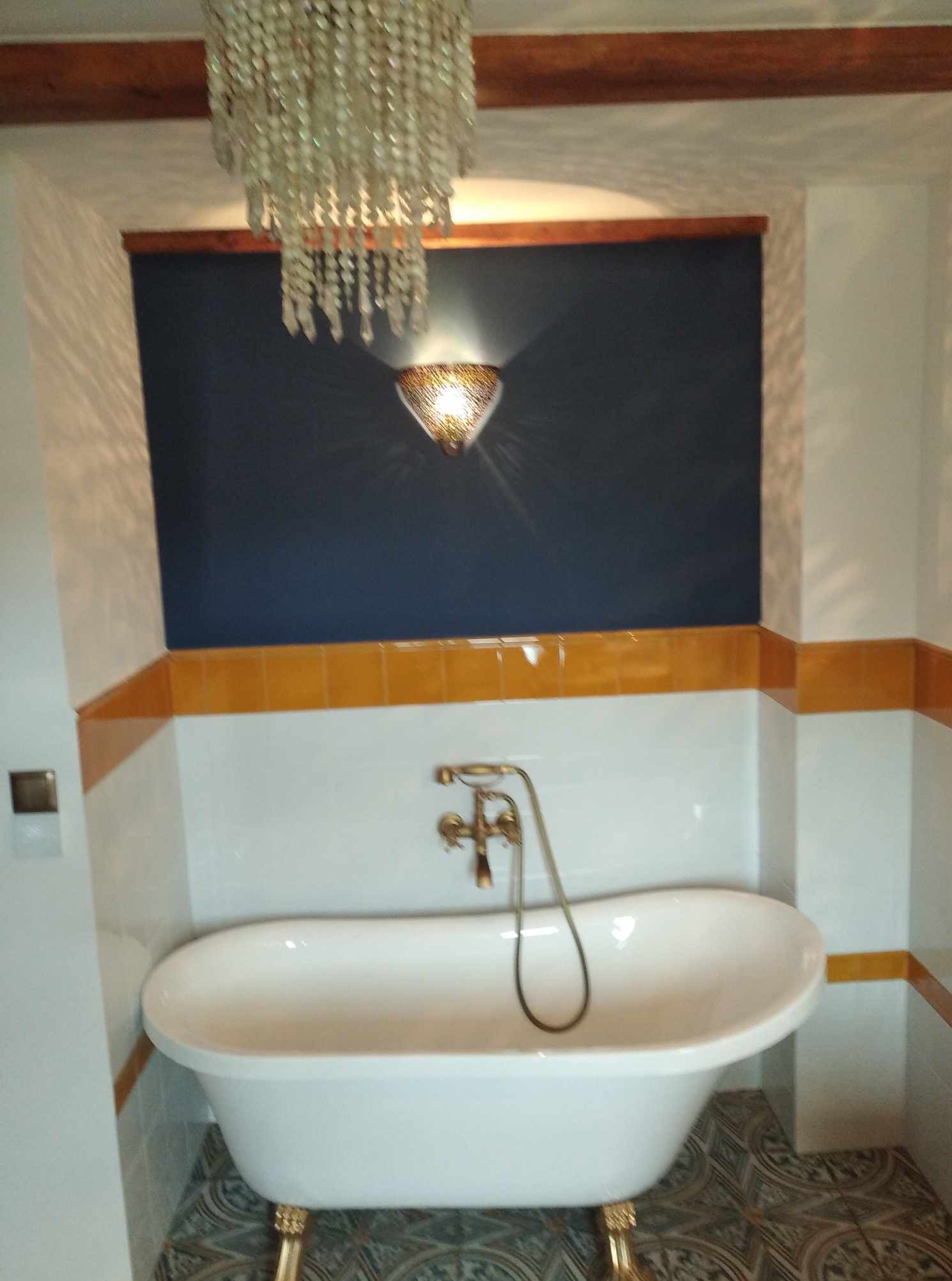 Biała wanna na złotych nóżkach w łazience z ciemnoniebieską ścianą, oświetlona ozdobnym żyrandolem i kinkietem. Żółte płytki stanowią akcent kolorystyczny.