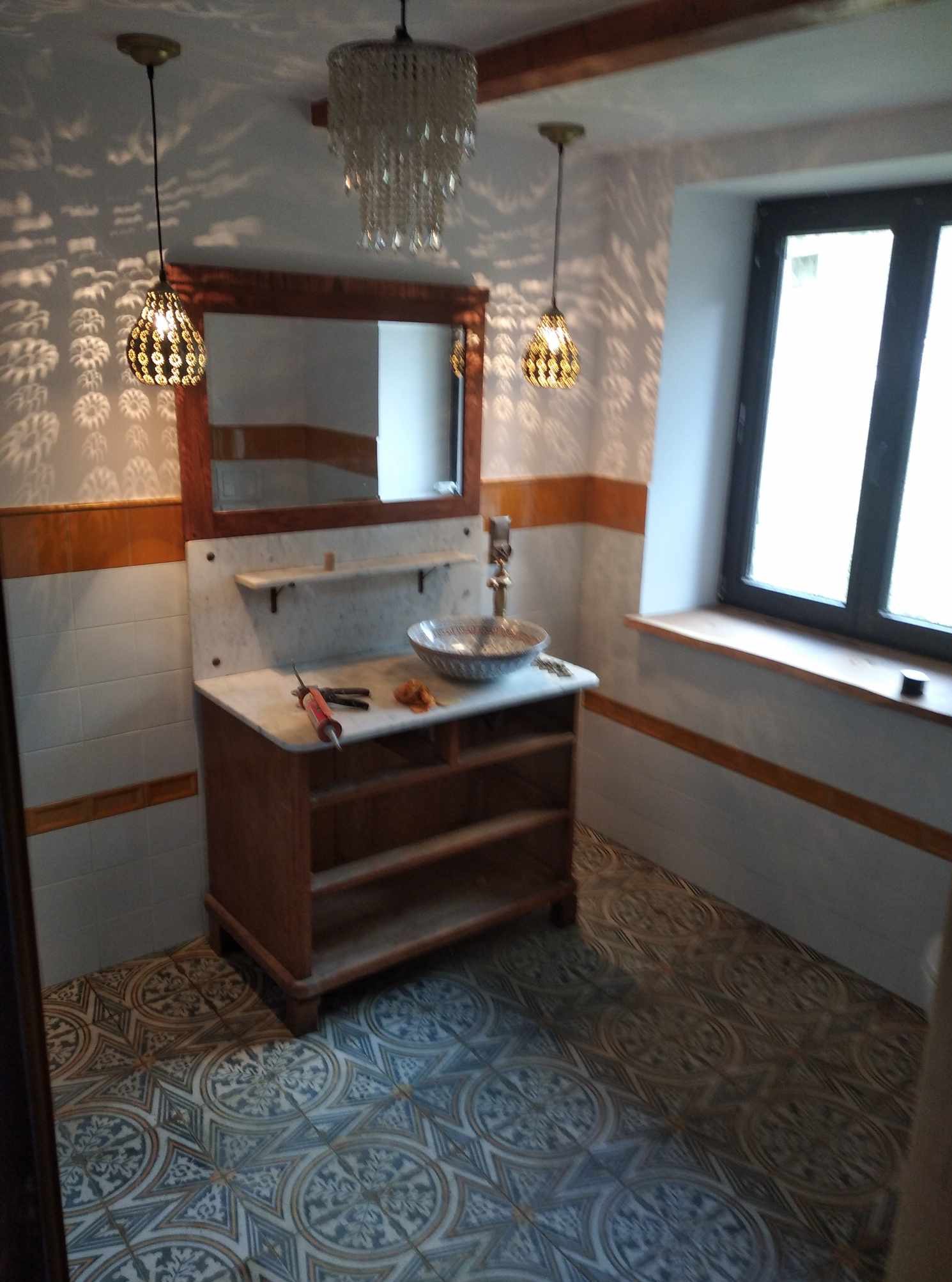 Wykończona łazienka z drewnianą szafką pod umywalką, wzorzystą podłogą i oknem. Widoczne lampy wiszące i akcesoria. Całość w stylu retro.