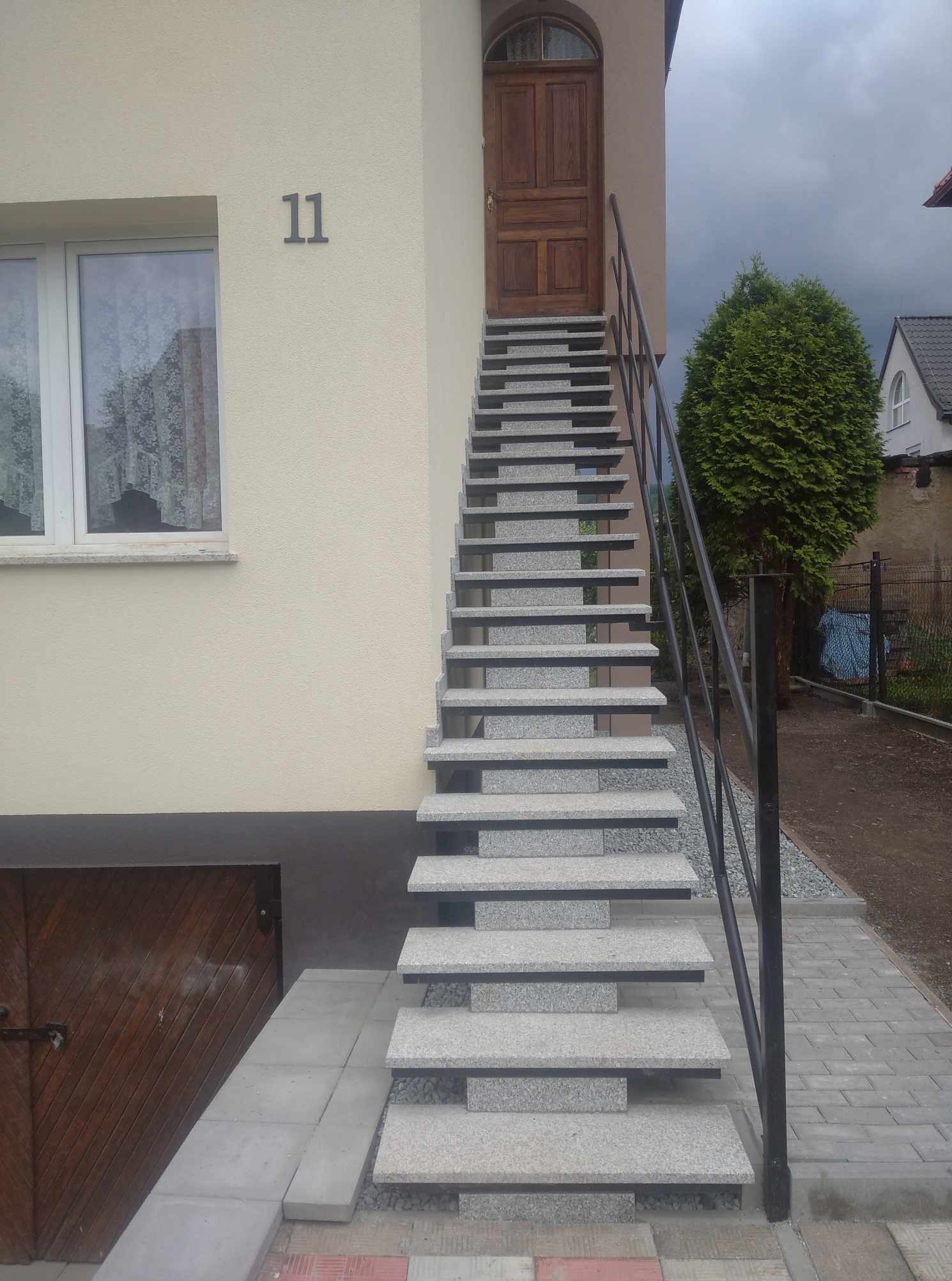 Zewnętrzne schody z granitu prowadzące do drewnianych drzwi wejściowych, z czarną metalową balustradą, numer domu '11' widoczny na elewacji.
