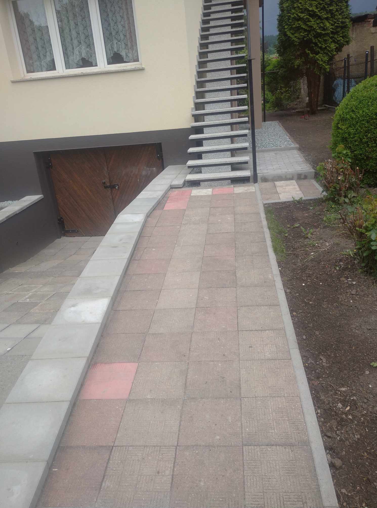 Ułożona kostka brukowa tworząca ścieżkę i podjazd do budynku z drewnianymi drzwiami garażowymi. Widoczne schody zewnętrzne z metalową barierką i roślinność.