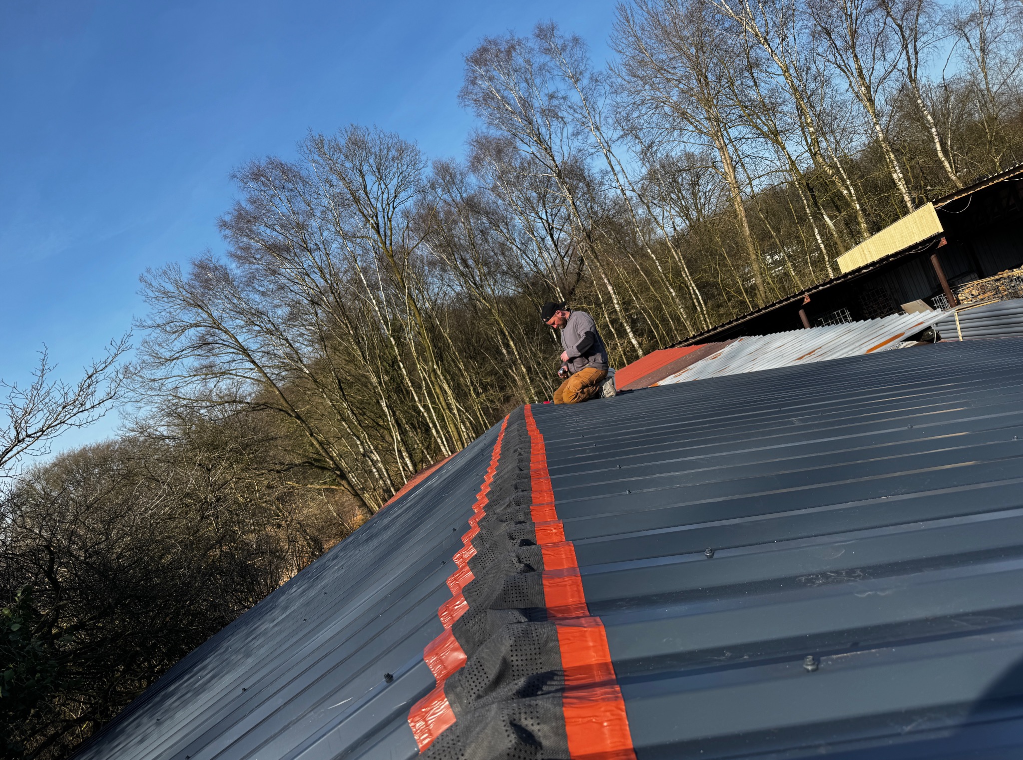 Pracownik montuje membranę dachową na szarym dachu z blachy na tle drzew i błękitnego nieba. Widok z góry, skupienie na detalu łączenia membrany.