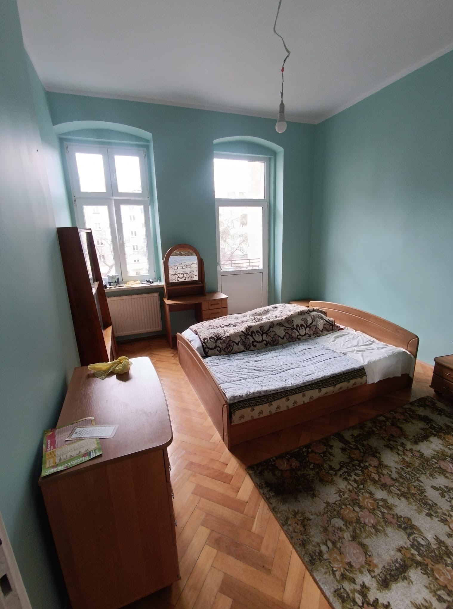 Pokój z łóżkiem, komodą i toaletką w odcieniach błękitu i brązu. Parkiet na podłodze, okna z białymi ramami. W tle drzwi balkonowe i wisząca żarówka.