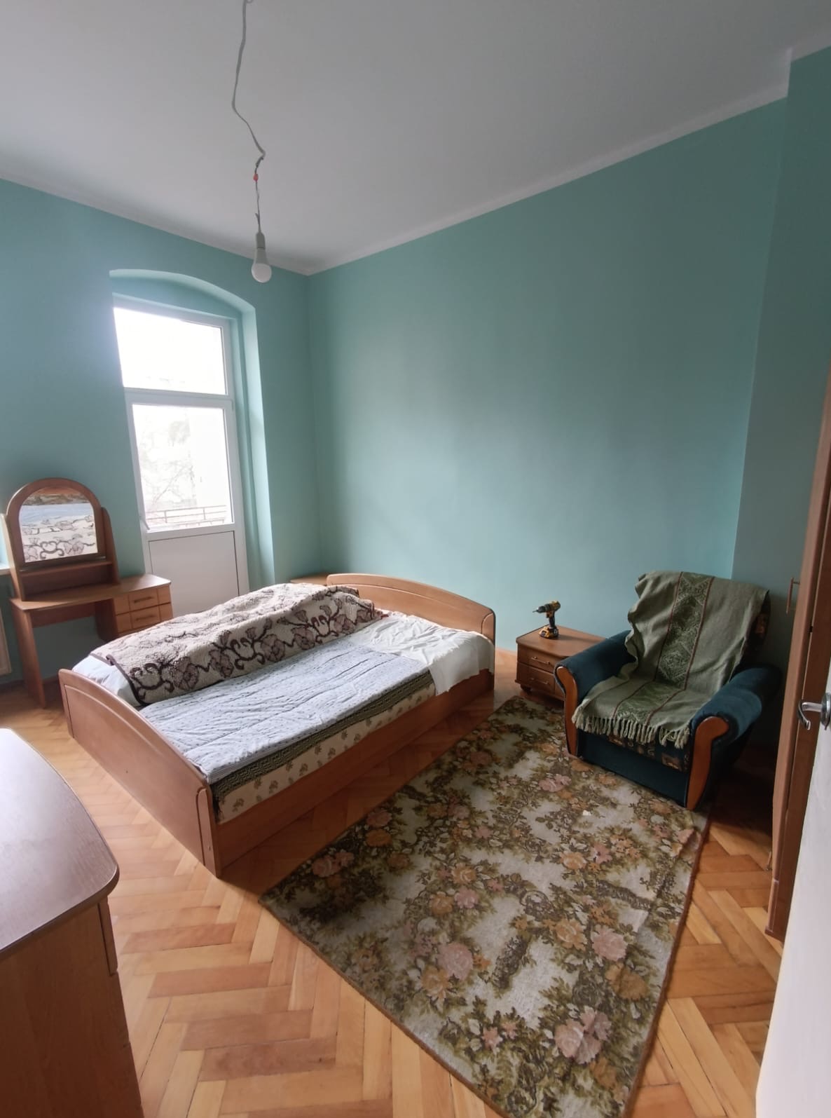 Pokój z łóżkiem, fotelem i toaletką, ściany pomalowane na błękitno. Na szafce nocnej leży wiertarka. Parkiet ułożony w jodełkę.