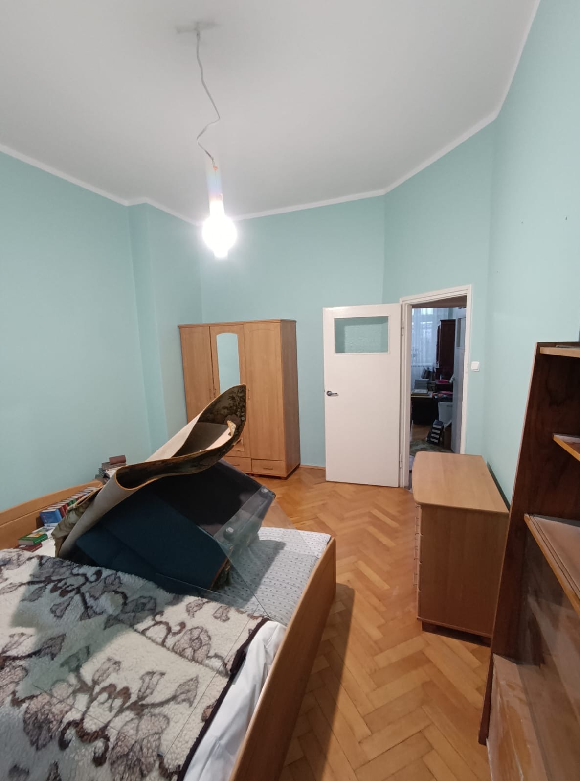 Pokój przed remontem: parkiet, błękitne ściany, drewniane meble, łóżko z narzutą i lampa. Widok na otwarte drzwi do kolejnego pomieszczenia.