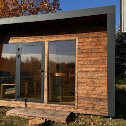 ORILL GROUP - Gotowa, drewniana sauna modułowa z przeszklonymi drzwiami, na trawie, z widocznym piecem i meblami w środku. Nietypowy kształt bryły.