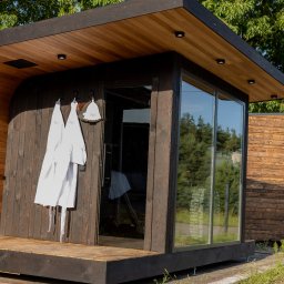 ORILL GROUP - Drewniana sauna modułowa z dużym oknem, na zewnątrz wiszą dwa białe szlafroki. Widoczne elementy wykończenia z ciemnego drewna i oświetlenie.
