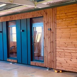 ORILL GROUP - Elewacja modułowego domu z drewnianą boazerią i ciemnymi, przeszklonymi drzwiami. Widoczne elementy konstrukcyjne i logo firmy na dole po prawej.