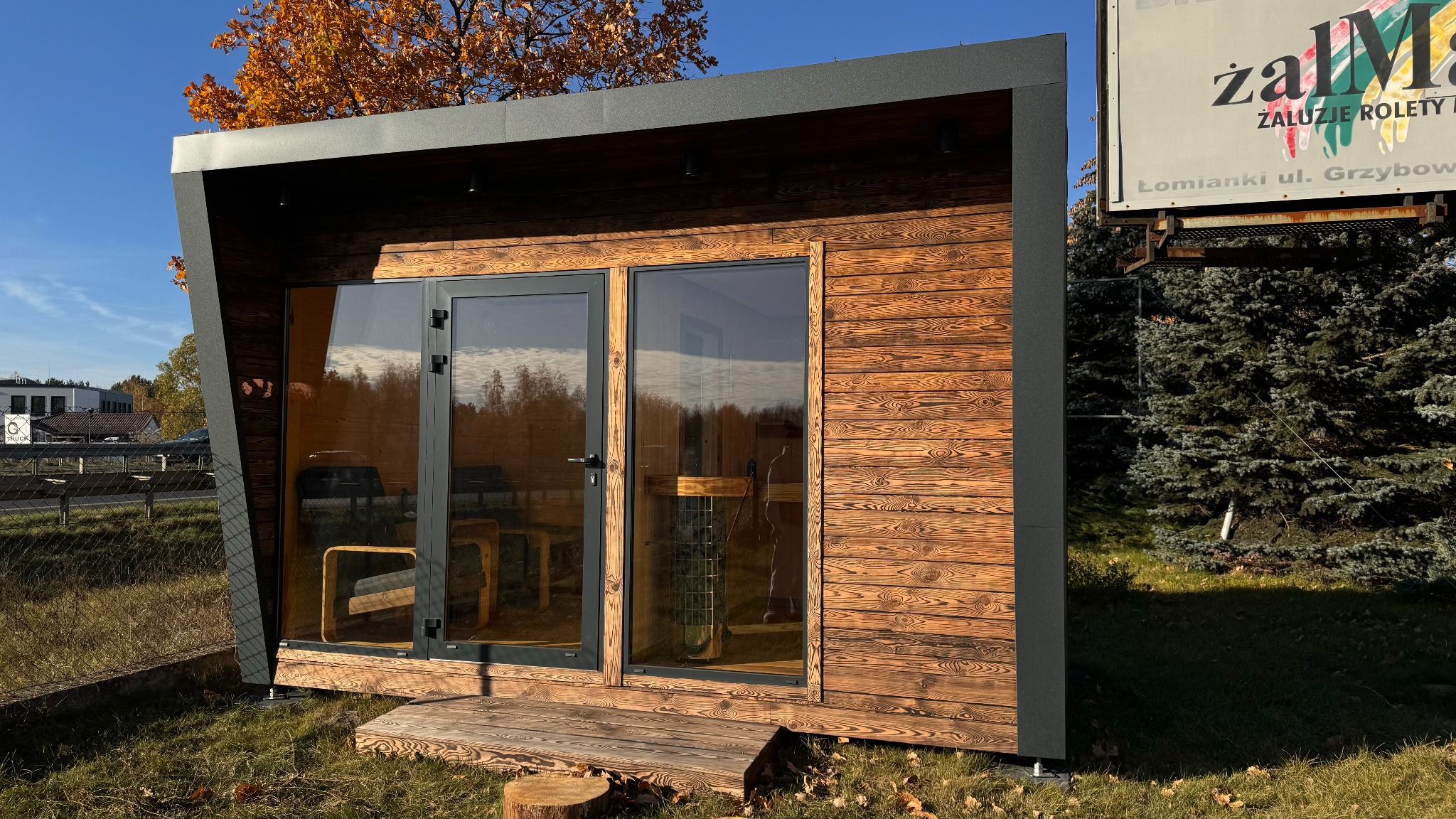 Gotowa, drewniana sauna modułowa z przeszklonymi drzwiami, na trawie, z widocznym piecem i meblami w środku. Nietypowy kształt bryły.
