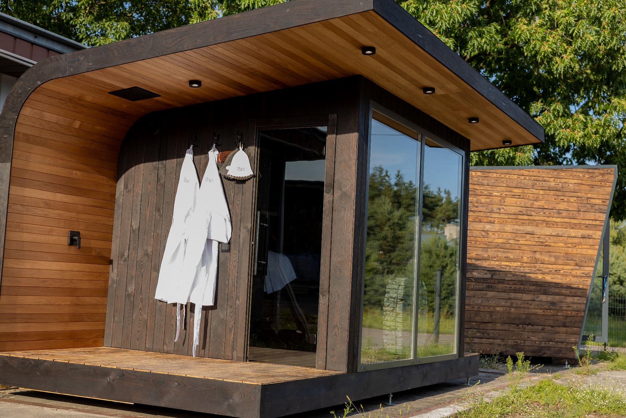 Drewniana sauna modułowa z dużym oknem, na zewnątrz wiszą dwa białe szlafroki. Widoczne elementy wykończenia z ciemnego drewna i oświetlenie.