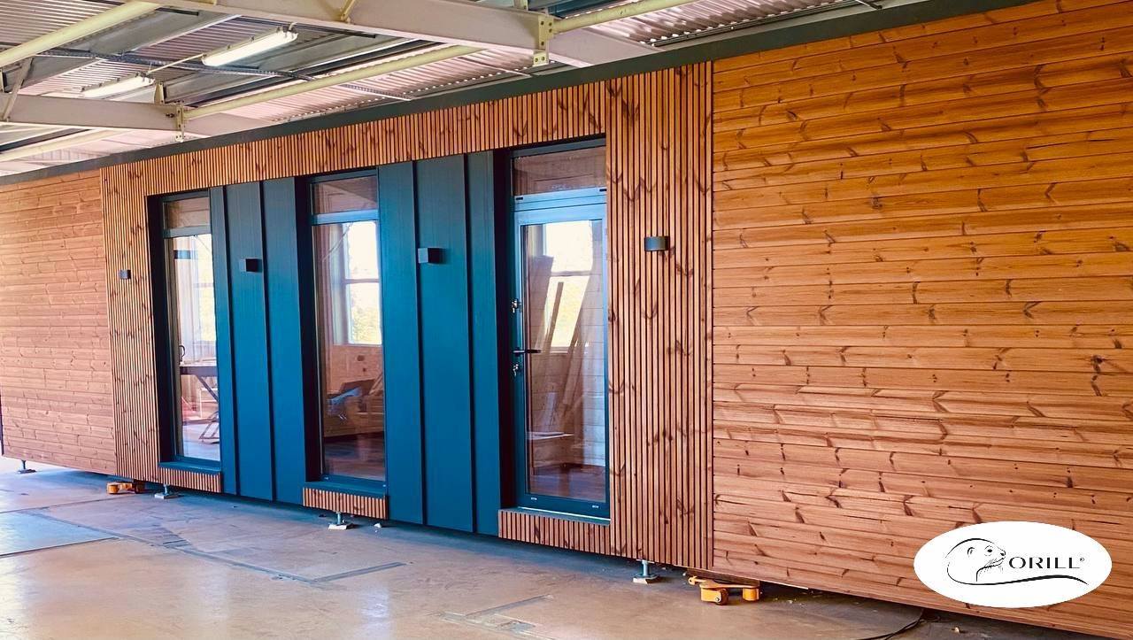 Elewacja modułowego domu z drewnianą boazerią i ciemnymi, przeszklonymi drzwiami. Widoczne elementy konstrukcyjne i logo firmy na dole po prawej.