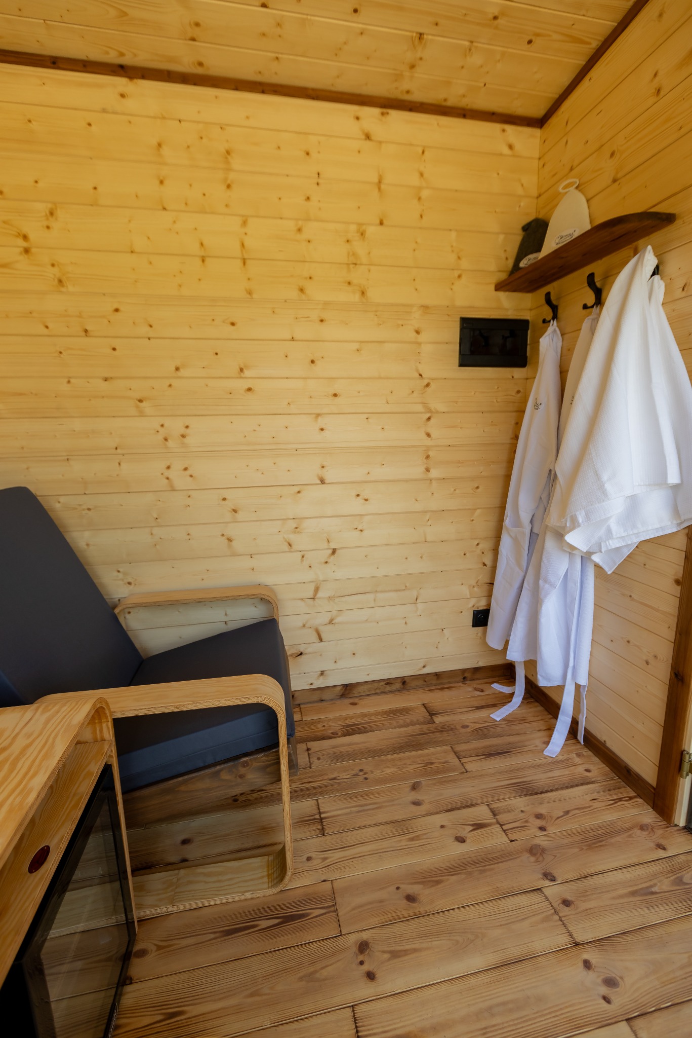 Wnętrze domku z drewna: fotel z szarą tapicerką, drewniany stolik z lodówką, wieszak z białymi szlafrokami, drewniane ściany i podłoga z desek.