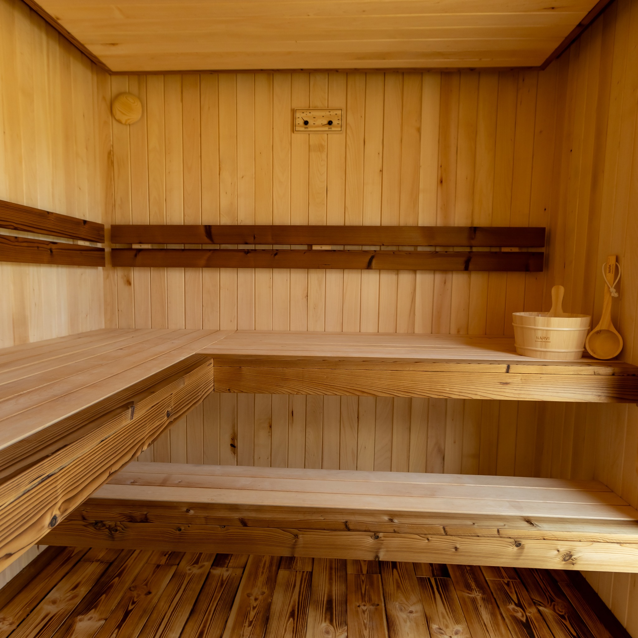 Wnętrze sauny z jasnego drewna, widoczne ławy, oparcia, termometr i akcesoria: wiadro oraz chochla marki Narvi. Ciepłe oświetlenie podkreśla strukturę drewna.