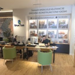 Drewno Dla Domu - Wnętrze biura z drewnianymi biurkami i zielonymi krzesłami. Na ścianie baner z ofertą tarasów, pergoli i elewacji. Pracownicy przy komputerach.