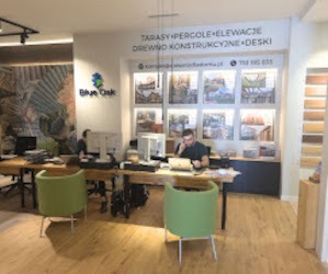 Wnętrze biura z drewnianymi biurkami i zielonymi krzesłami. Na ścianie baner z ofertą tarasów, pergoli i elewacji. Pracownicy przy komputerach.