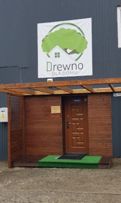 Wejście do składu drewna z drewnianym zadaszeniem i szyldem 'Drewno dla domu' z zielonym drzewem na białym tle. Widok na brązowe drzwi wejściowe i fragment elewacji.