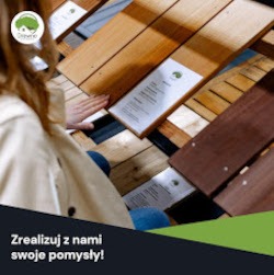 Kobieta dotyka dłonią próbki desek tarasowych o różnych odcieniach brązu, umieszczonych na ekspozytorze w sklepie. Widoczny fragment reklamy.