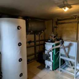 Nowoczesny piec na paliwo stałe z podajnikiem, zbiornik akumulacyjny i instalacja CO w kotłowni z beżowo-szarą terakotą. Widoczne rury i zawory.