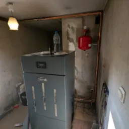 Szary piec Ferroli z podłączonymi miedzianymi rurami, czerwonym zbiornikiem wyrównawczym i butelką wody na górze, w surowym wnętrzu kotłowni.