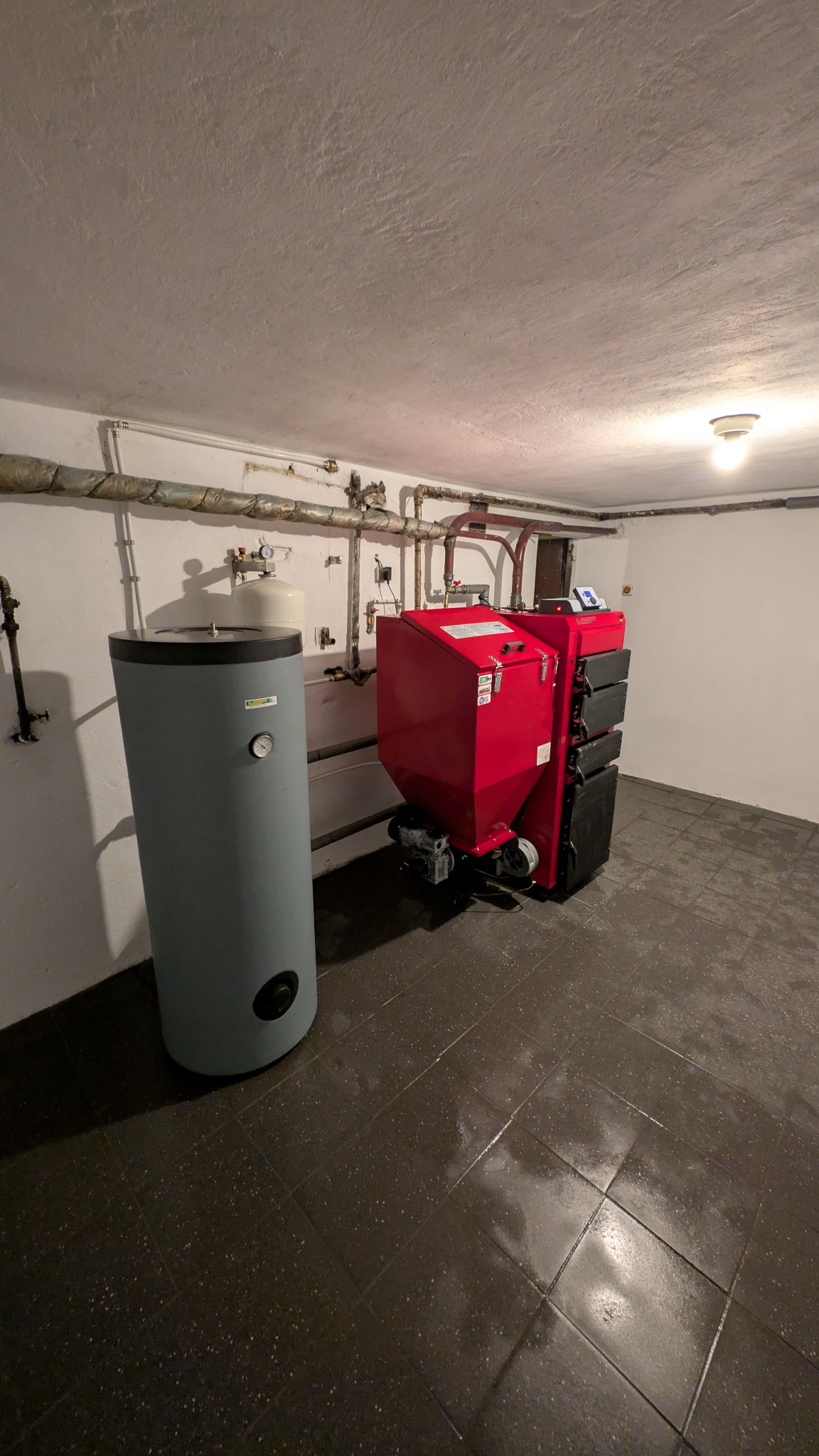 Czerwony piec na paliwo stałe z zasobnikiem i szary zasobnik CWU w kotłowni. Widoczne rury instalacji grzewczej pod sufitem.