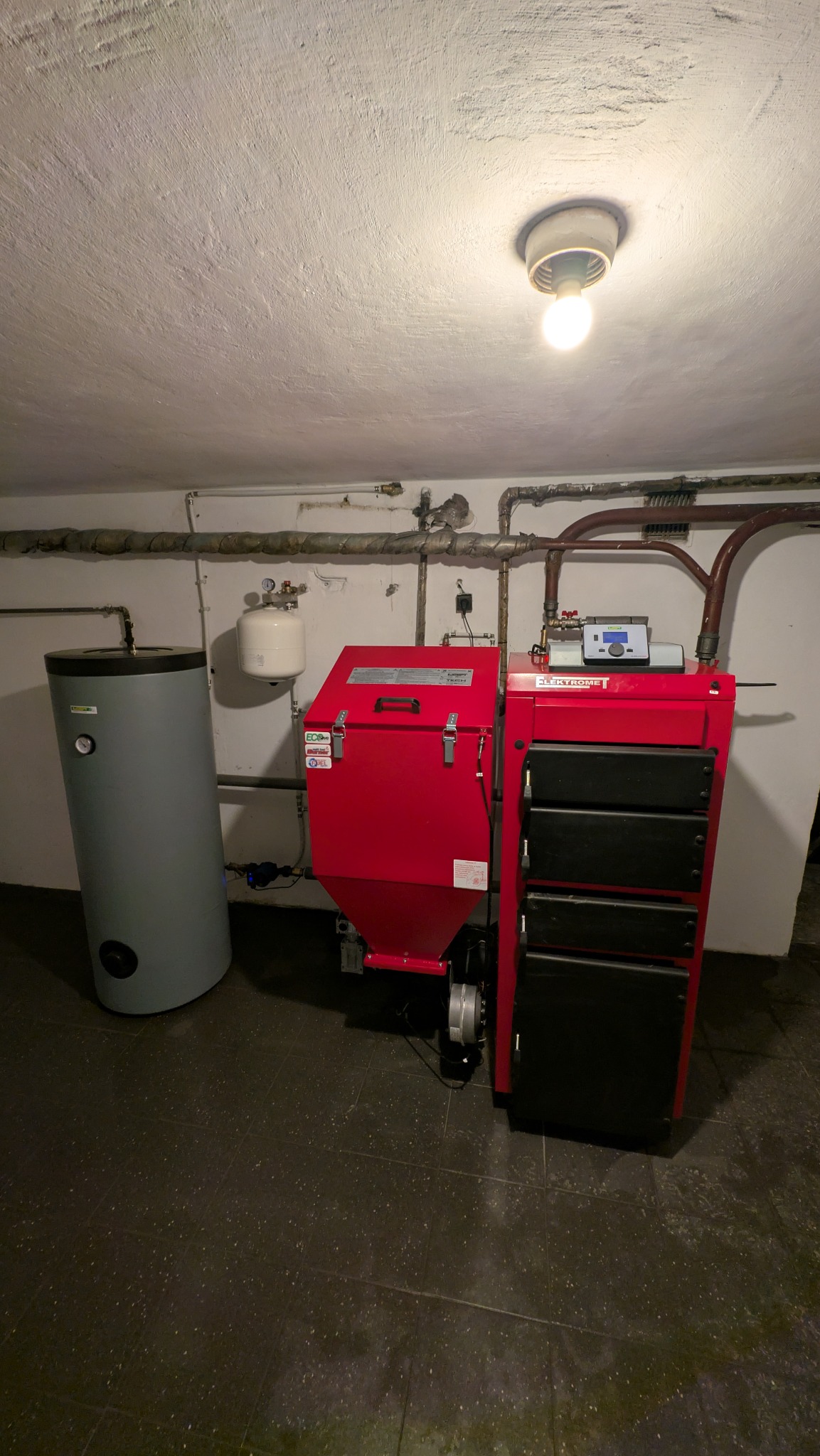 Czerwony piec na paliwo stałe Elektromet z zasobnikiem, szary bojler, rury instalacji grzewczej na tle ściany i podłogi wyłożonej ciemnymi płytkami w piwnicy.