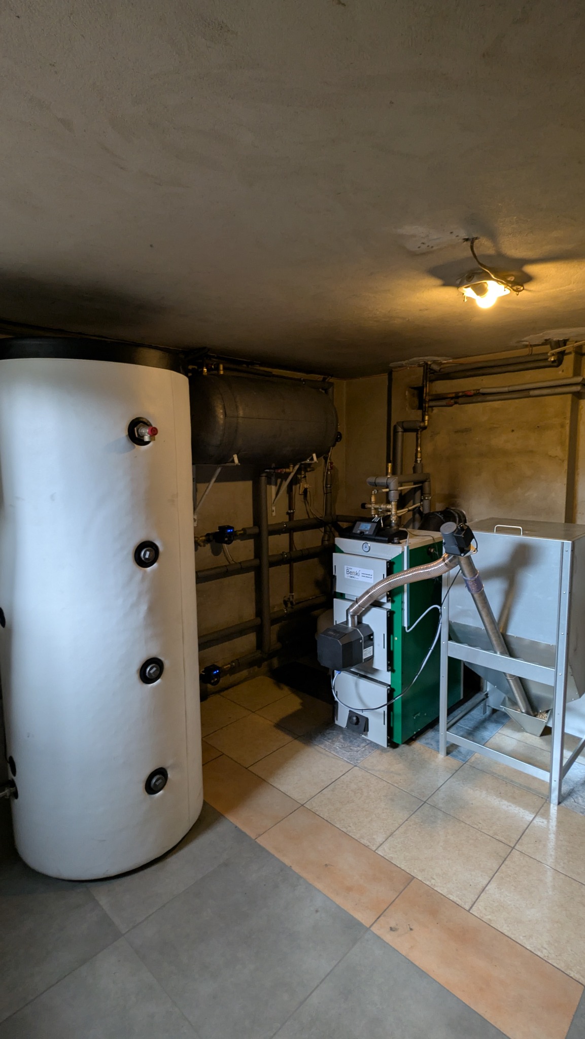 Nowoczesny piec na paliwo stałe z podajnikiem, zbiornik akumulacyjny i instalacja CO w kotłowni z beżowo-szarą terakotą. Widoczne rury i zawory.