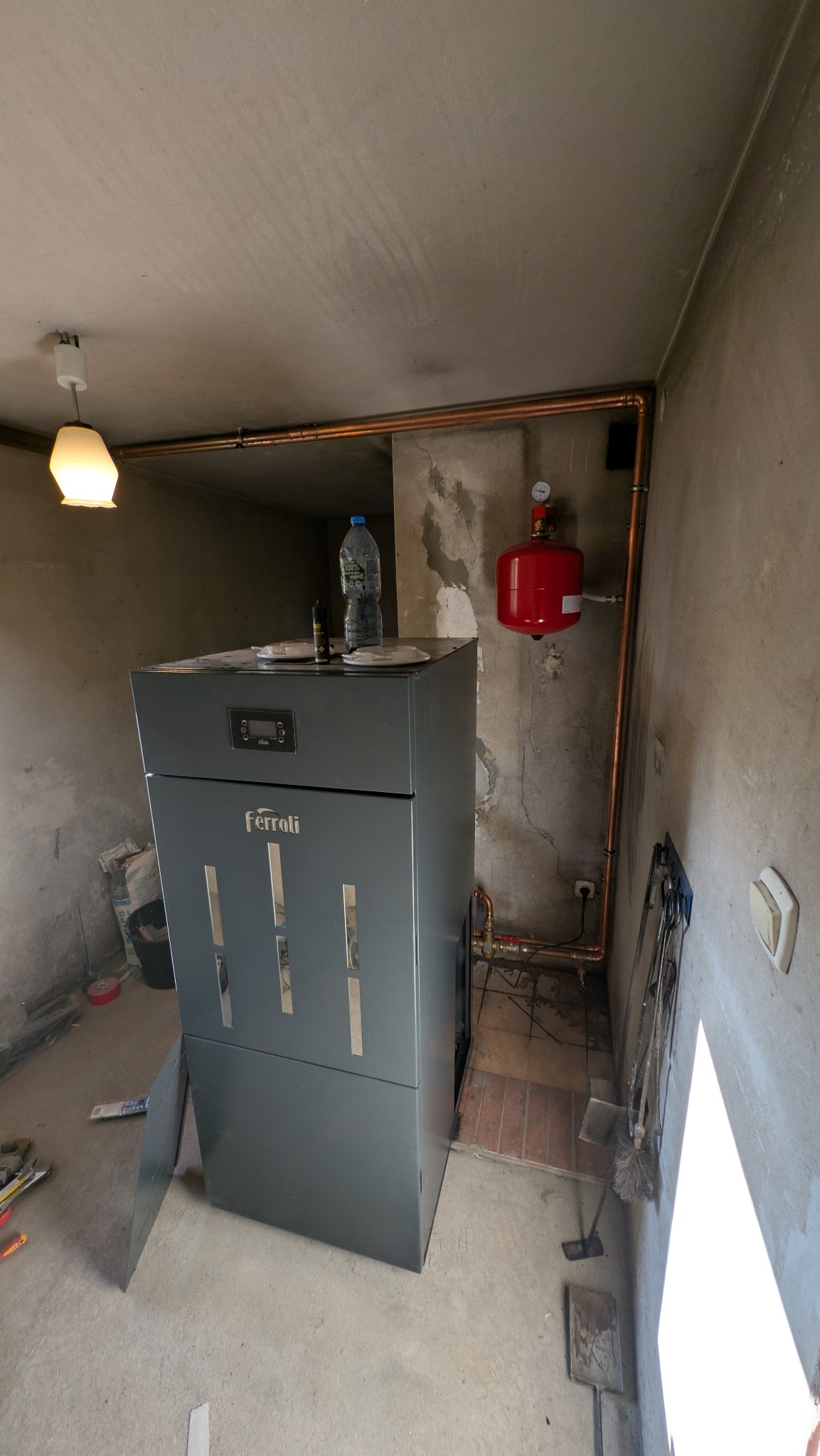 Szary piec Ferroli z podłączonymi miedzianymi rurami, czerwonym zbiornikiem wyrównawczym i butelką wody na górze, w surowym wnętrzu kotłowni.