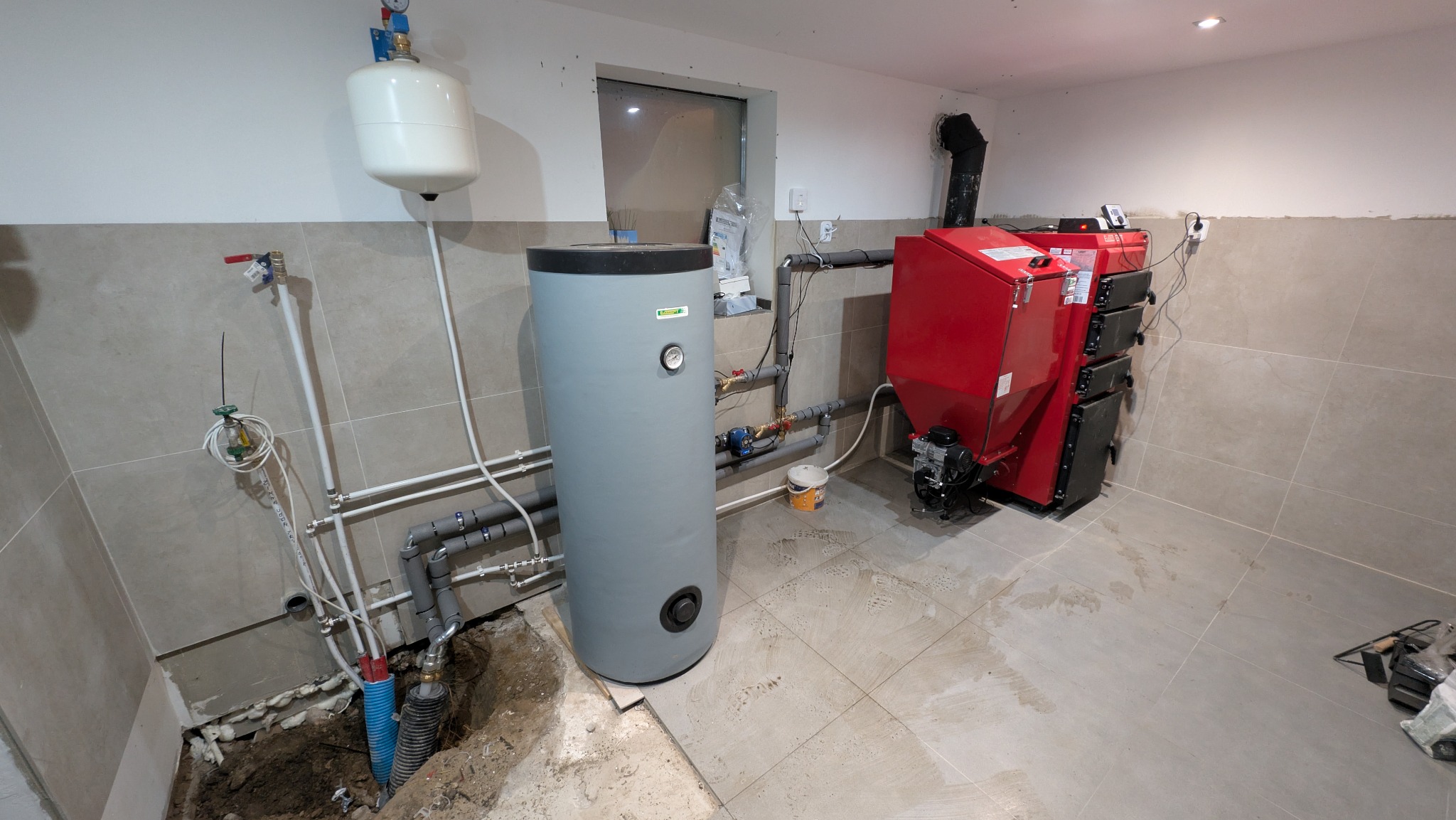 Czerwony piec na paliwo stałe z szarym zbiornikiem buforowym w kotłowni, widoczne rury instalacji i wykop w podłodze. Realizacja z jasnymi płytkami na ścianach.