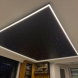 Andrei Malyshka Sufity napinane Pawbud - Nowoczesny sufit z efektem gwieździstego nieba, obramowany listwami LED. Widok na część umeblowanego wnętrza z żaluzjami i przeszkloną witryną.