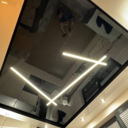 Andrei Malyshka Sufity napinane Pawbud - Błyszcząca, czarna powierzchnia sufitu z wbudowanym oświetleniem LED w kształcie krzyża, odbijająca wnętrze pokoju. Widoczny fragment kuchni i okno z żaluzjami.