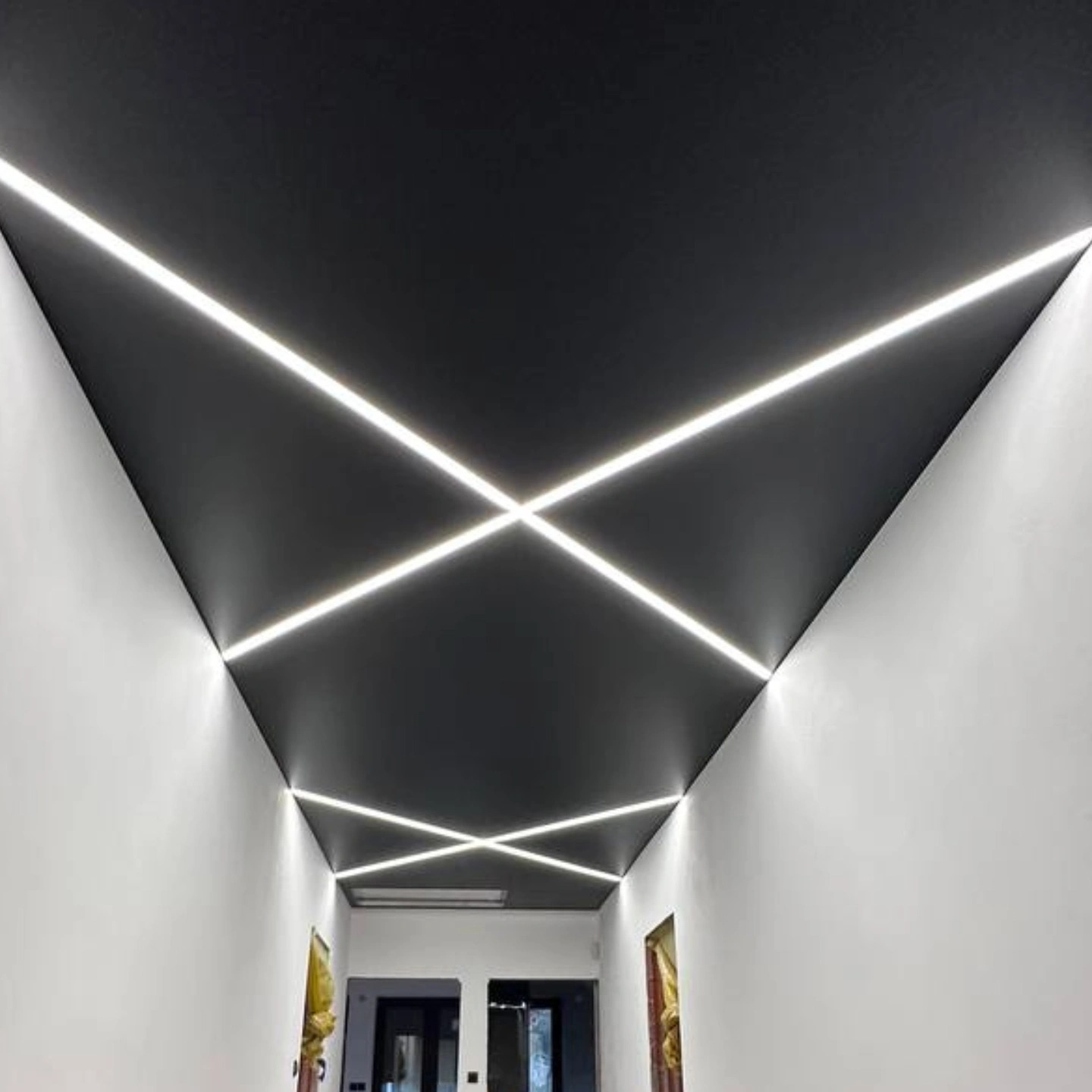 Nowoczesny, czarny sufit z krzyżującymi się liniami LED, tworzącymi geometryczny wzór. Białe ściany i drzwi w tle. Unikalne oświetlenie wnętrza.