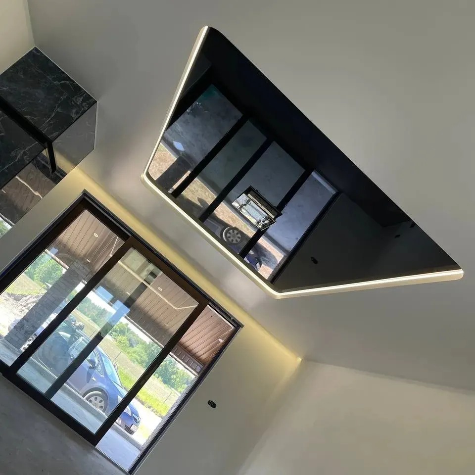 Nowoczesny pokój z oknem i podwieszanym sufitem z czarnym lustrzanym panelem i oświetleniem LED. W lustrze odbija się otoczenie.