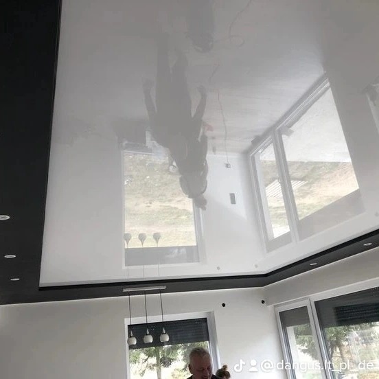 Biały, lśniący sufit z efektem lustrzanym, odbijający wnętrze pokoju z oknami i osobą. Nowoczesne wykończenie wnętrz.