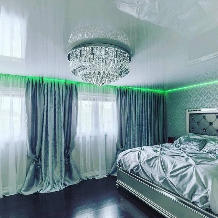 Błyszczący sufit z kryształowym żyrandolem w sypialni, podświetlony zielonym neonem. Eleganckie zasłony i łóżko z pikowanym zagłówkiem dopełniają luksusowego wnętrza.