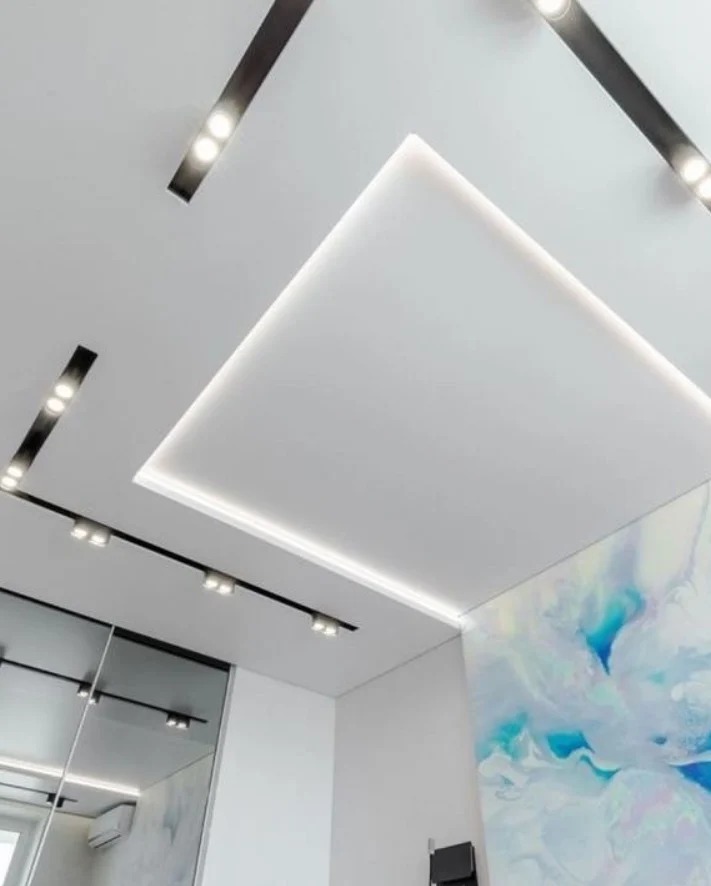 Biały sufit z podwieszaną, kwadratową strukturą i oświetleniem LED, uzupełniony czarnymi szynami z reflektorami punktowymi. W tle widoczna ściana z abstrakcyjnym wzorem.