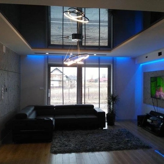 Nowoczesny salon z czarnym, lustrzanym sufitem napinanym, oświetleniem LED i designerską lampą wiszącą. Widok na okno z żaluzjami i czarną sofę.